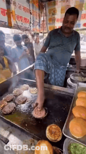 A Man Is Cooking Burgers On A Grill With His Foot GIF