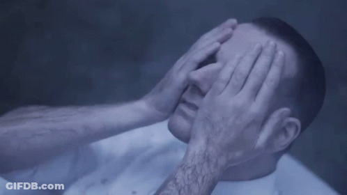 A Man Is Covering His Eyes With His Hands While Laying On A Bed . GIF