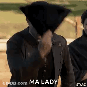 A Man Is Covering His Face With A Hat While Standing Next To Another Man . GIF