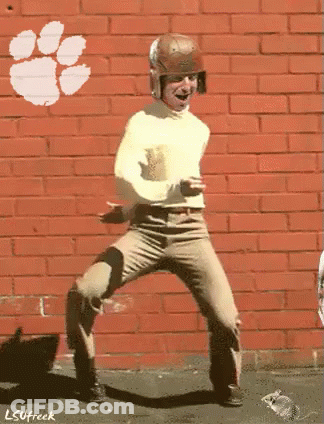 A Man Is Dancing In Front Of A Brick Wall That Has A Clemson Logo On It GIF