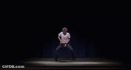 A Man Is Dancing On A Stage In A Dark Room With A Blue Curtain Behind Him . GIF