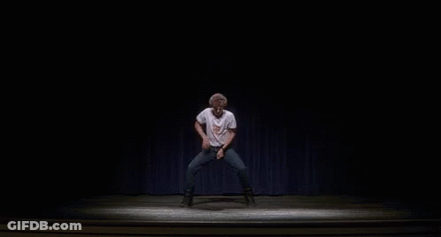 A Man Is Dancing On A Stage In A Dark Room With A Blue Curtain Behind Him . GIF