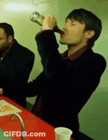 A Man Is Drinking From A Bottle While Sitting At A Table With Another Man . GIF