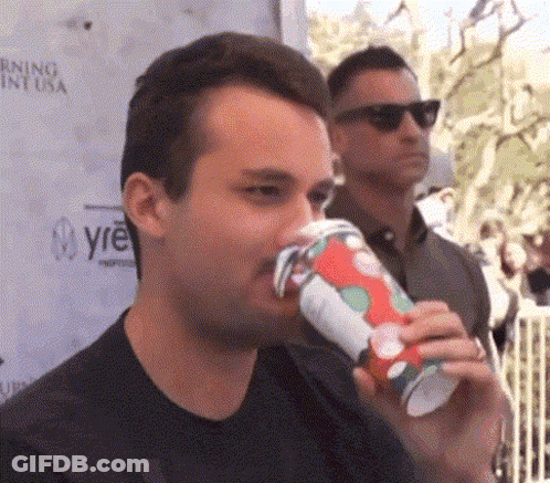 A Man Is Drinking From A Red And White Cup While Another Man Stands Behind Him . GIF