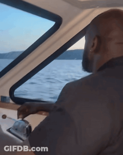 A Man Is Driving A Boat In The Water And Looking Out The Window GIF