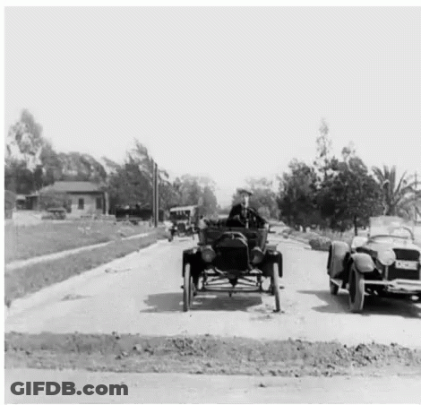 A Man Is Driving A Car Down A Street Next To A Convertible Car . GIF
