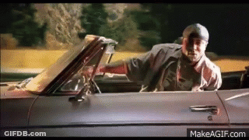 A Man Is Driving A Convertible Car On A Road . GIF