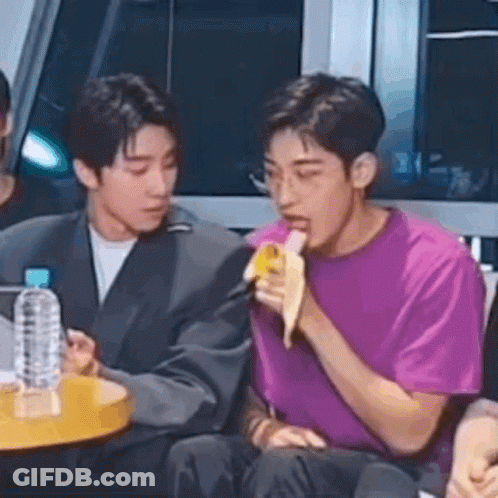 A Man Is Eating A Banana In Front Of A Microphone While Sitting Next To Another Man . GIF