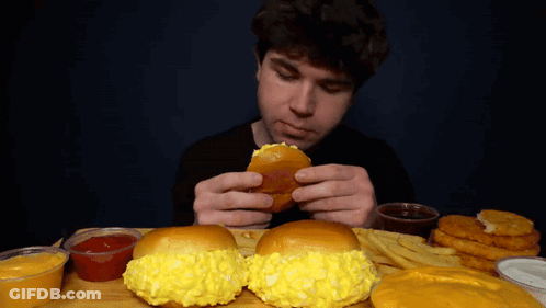 A Man Is Eating A Hamburger And French Fries At A Table GIF