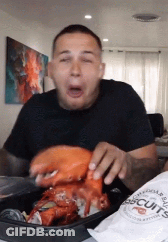 A Man Is Eating A Large Lobster With A Bag Of Biscuits In Front Of Him GIF