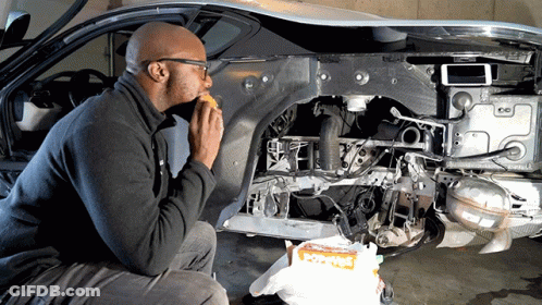 A Man Is Eating A Sandwich In Front Of A Car That Is Being Built GIF