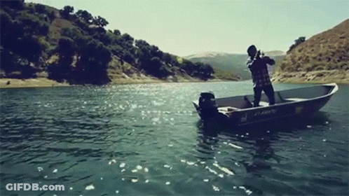 A Man Is Fishing In A Boat On A Lake With Mountains In The Background . GIF