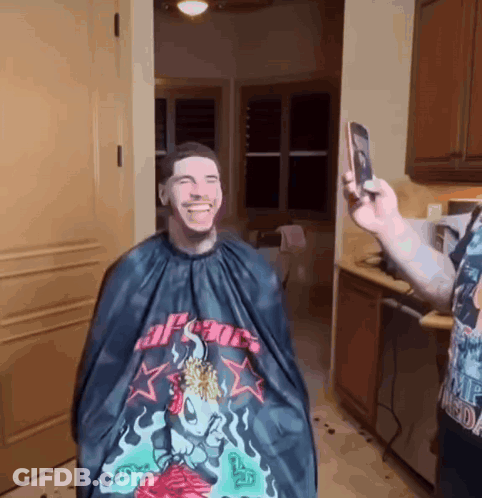 A Man Is Getting His Hair Cut By A Man Wearing A Cape That Says Apsoc On It GIF