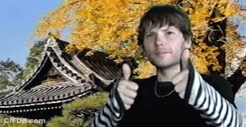 A Man Is Giving A Thumbs Up In Front Of A Tree And A Building GIF