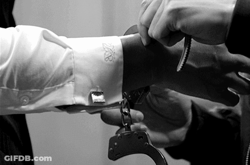 A Man Is Handcuffed To Another Man 'S Arm With A White Shirt With The Letter B On The Sleeve GIF