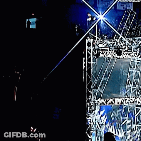 A Man Is Hanging From A Rope In A Dark Room With A Sign That Says ' Wwe ' On It . GIF