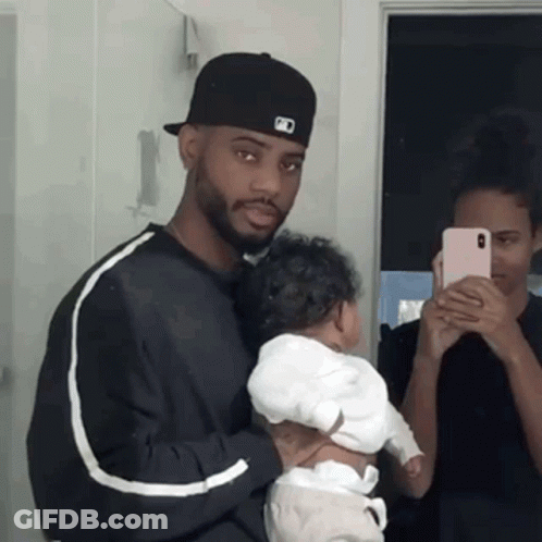 A Man Is Holding A Baby In Front Of A Mirror While A Woman Takes A Picture Of Them . GIF