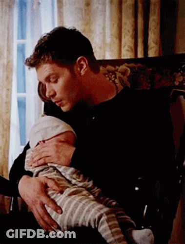 A Man Is Holding A Baby In His Lap While Sitting In A Chair . GIF