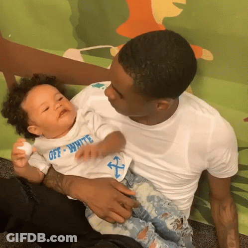 A Man Is Holding A Baby Who Is Wearing A White Shirt That Says Off White GIF
