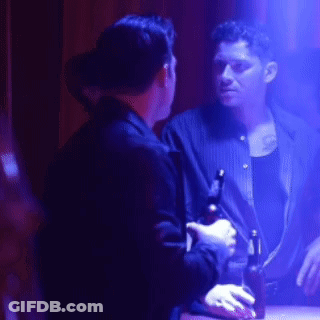 A Man Is Holding A Bottle Of Beer And Talking To Another Man In A Dark Room GIF