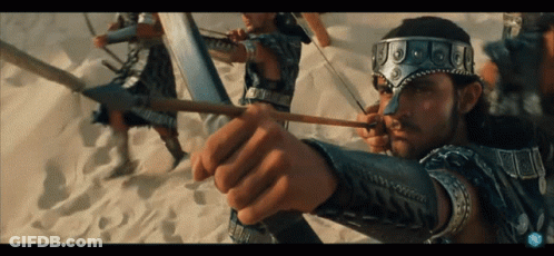 A Man Is Holding A Bow And Arrow With A Few Other Men In The Background GIF