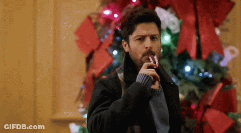 A Man Is Holding A Candy Cane In His Mouth While Standing In Front Of A Christmas Tree . GIF