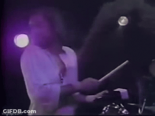 A Man Is Holding A Drum Stick In His Hand On A Stage . GIF