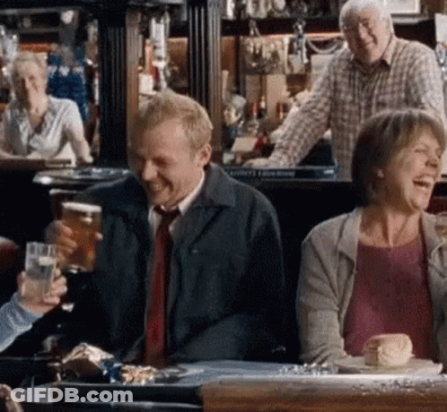 A Man Is Holding A Glass Of Beer While Sitting At A Table With Other People In A Bar . GIF