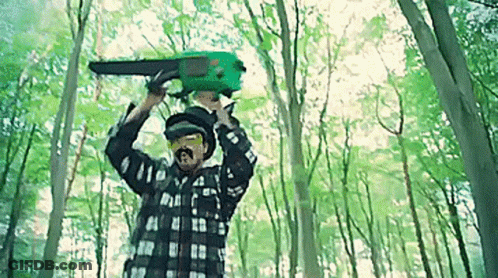 A Man Is Holding A Green Chainsaw Over His Head In The Woods . GIF