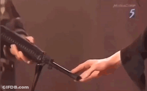 A Man Is Holding A Gun In Front Of A Man Standing Next To A Man Holding A Gun . GIF