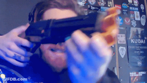 A Man Is Holding A Gun In Front Of A Wall With Stickers On It Including One That Says Kudai GIF