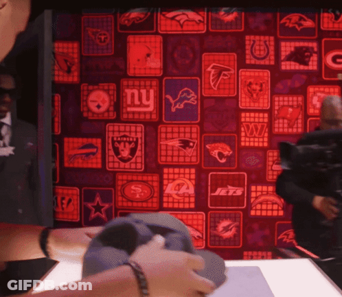 A Man Is Holding A Hat In Front Of A Wall With Logos For The New York Jets And New York Raiders GIF