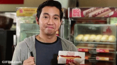 A Man Is Holding A Hot Dog In Front Of A Big Hot Dog Stand GIF