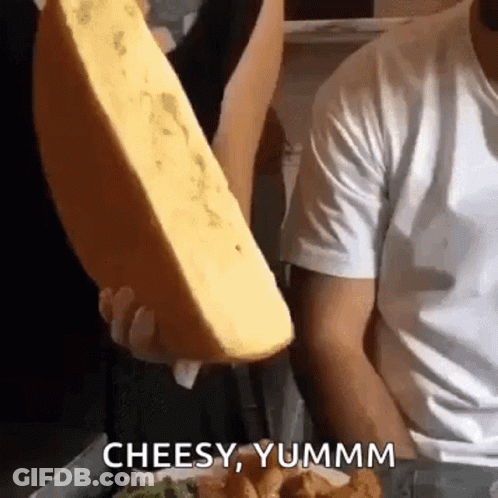 A Man Is Holding A Large Piece Of Cheese And Saying `` Cheesy Yummm '' . GIF
