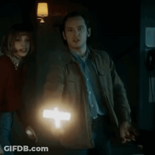 A Man Is Holding A Lighted Cross In Front Of A Woman In A Dark Room . GIF