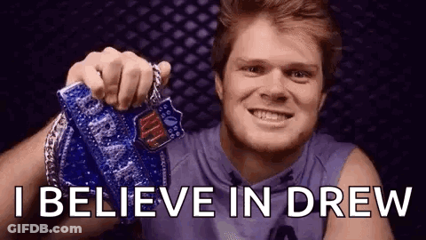 A Man Is Holding A Medal In His Hand And Smiling With The Words `` I Believe In Drew '' Behind Him . GIF