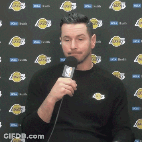 A Man Is Holding A Microphone In Front Of A Wall With Lakers Logos GIF