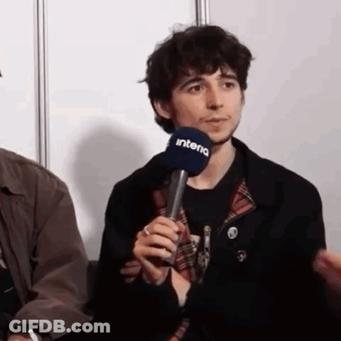 A Man Is Holding A Microphone In His Hand While Sitting In Front Of A Wall . GIF