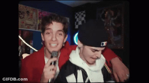 A Man Is Holding A Microphone Next To Another Man In A Hat . GIF