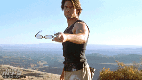 A Man Is Holding A Pair Of Glasses In Front Of A Poster For Mission Impossible 2 GIF