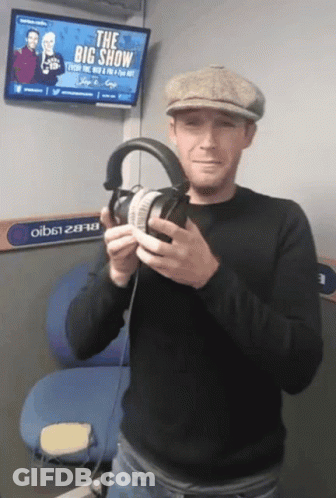 A Man Is Holding A Pair Of Headphones In Front Of A Big Show Sign GIF