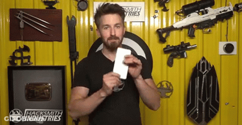 A Man Is Holding A Piece Of Paper In Front Of A Wall That Has Hacksmith Industries Written On It GIF