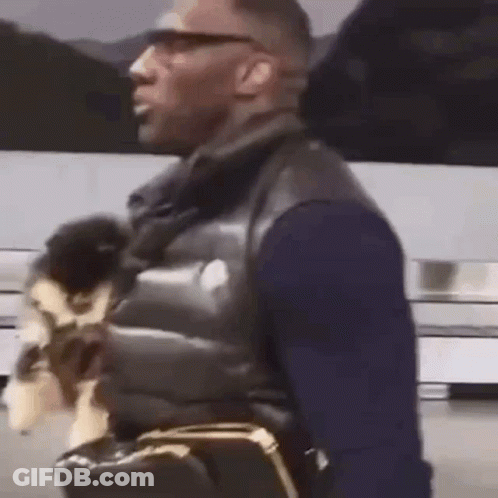 A Man Is Holding A Small Dog In His Arms While Walking Down The Street . GIF