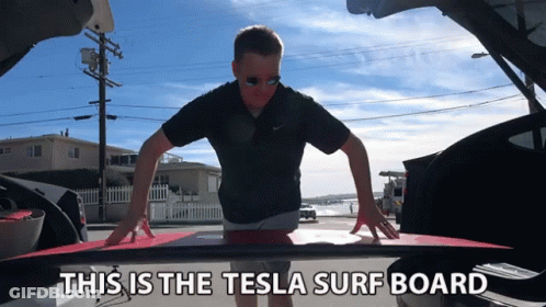 A Man Is Holding A Surfboard In The Back Of A Tesla Car GIF