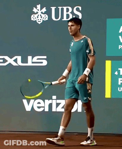 A Man Is Holding A Tennis Racquet In Front Of A Wall With Ubs And Verizon Advertisements GIF
