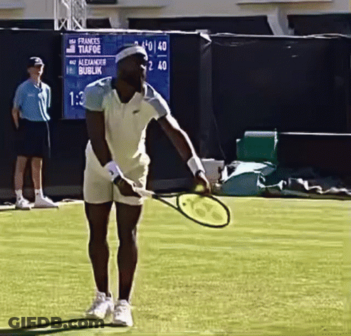A Man Is Holding A Tennis Racquet On A Court With A Scoreboard Behind Him That Says Frances Tiafoe GIF