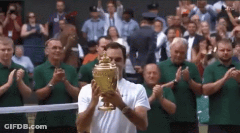 A Man Is Holding A Tennis Trophy In Front Of A Crowd . GIF
