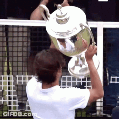 A Man Is Holding A Trophy Above His Head In Front Of A Tennis Net GIF