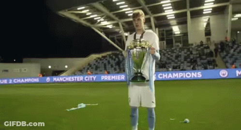 A Man Is Holding A Trophy On A Soccer Field With Manchester City Premier League Banners Behind Him GIF