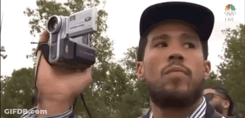 A Man Is Holding A Video Camera In His Hand And Looking At The Camera . GIF